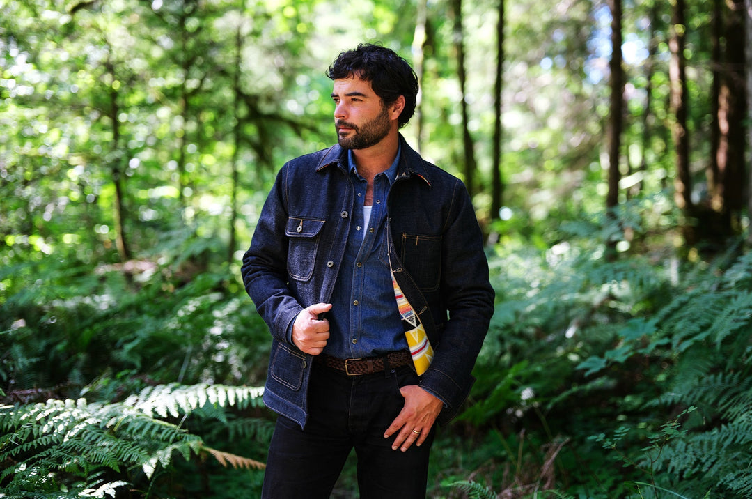 Man wearing the Ginew Bill Barn denim coat over a collared shirt, standing in a forest