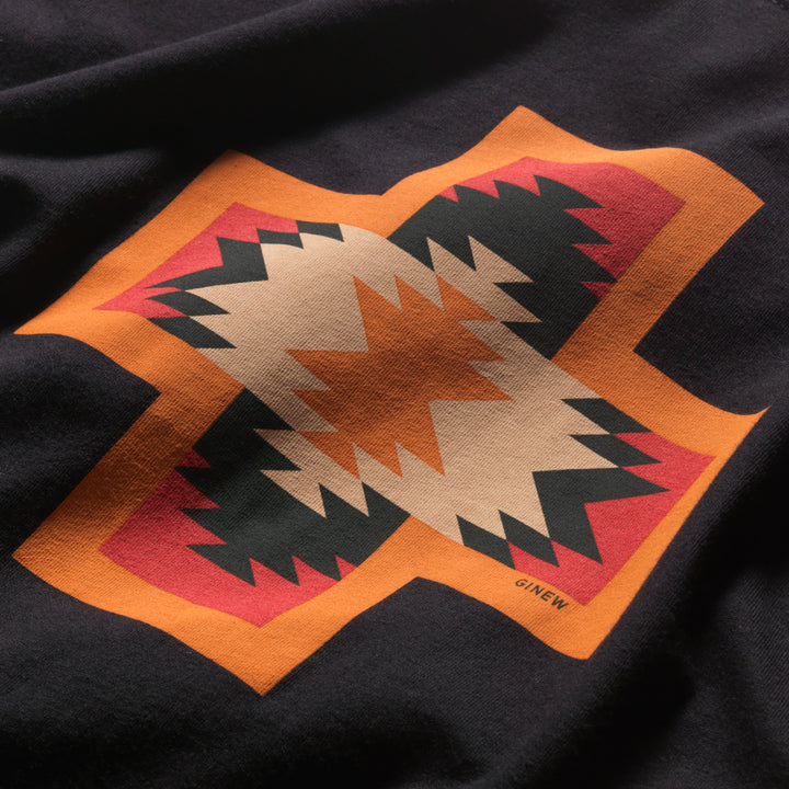 Close up of Made in USA black t-shirt with Native American Four Direction symbol on front in orange, red, tan and black with word Ginew 