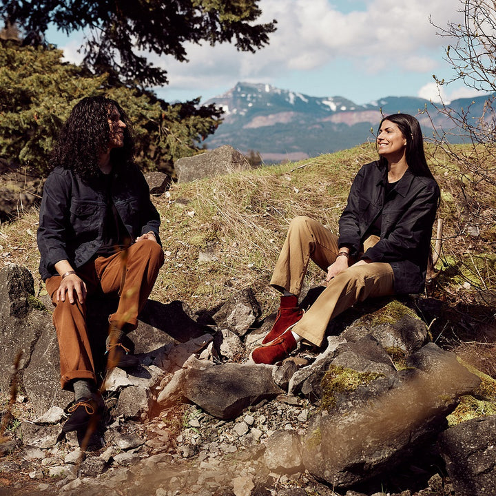Two models sitting on rocks with a Mountain View behind them. Male model on left wearing Superior Pant in Adobe and Black Wax Canvas Jacket. Female model on right wearing Superior Pant in Khaki and Black Wax Canvas Jacket.