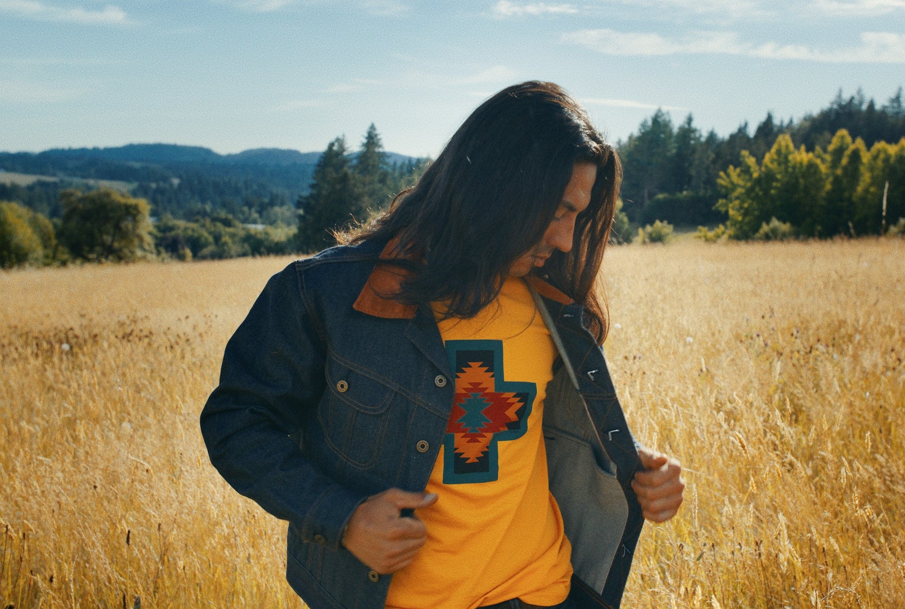 Native American male model wearing Ginew selvedge denim jacket and yellow made in USA tshirt standing in wheat field. 