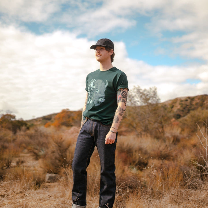 Man in canyon with Green bison t-shirt and selvedge denim jean and black wax canvas hat 