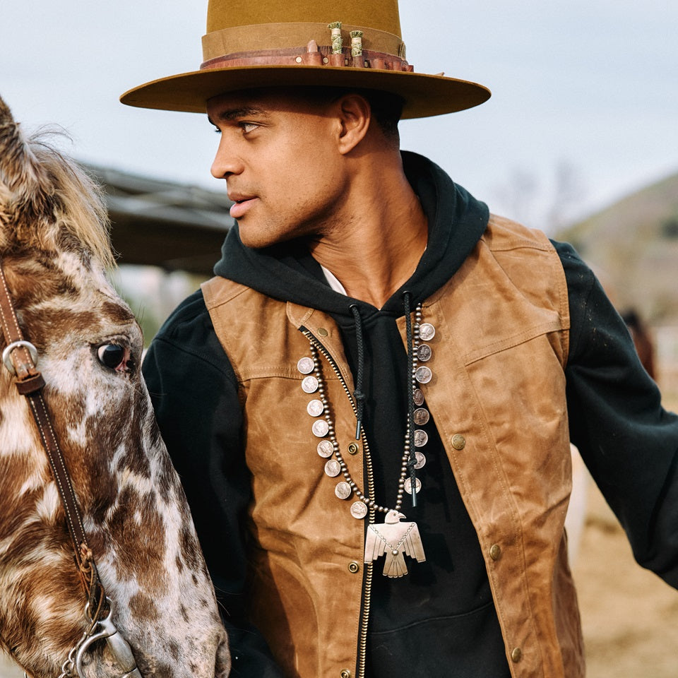 Native American Ginew brown wax canvas vest on model with horse. Custom hat by Ginew + Thundervoice hat co in tan brown.