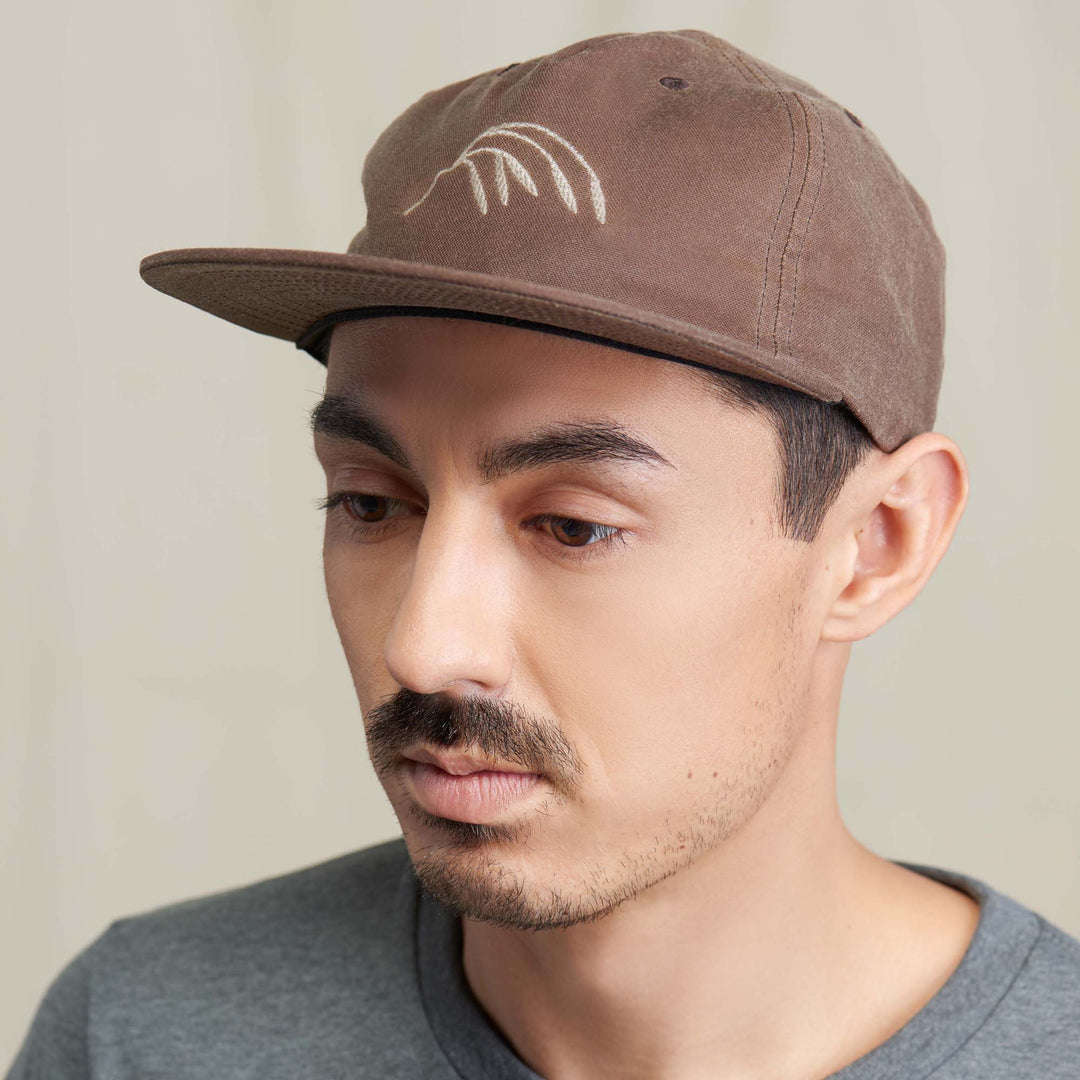 A light brown waxed canvas ball cap featuring a Crow Wing stitching in beige thread at the center front panel on a male model wearing a grey shirt in front of a neutral background.