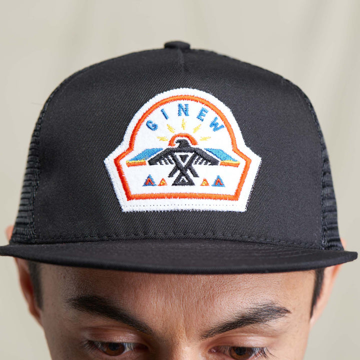 A male model wears the black Trucker Hat with a white Ginew patch featuring a thunderbird design stiched in blue, black, red, yellow, and orange thread. The model wears a grey shirt and poses against a neutral background.