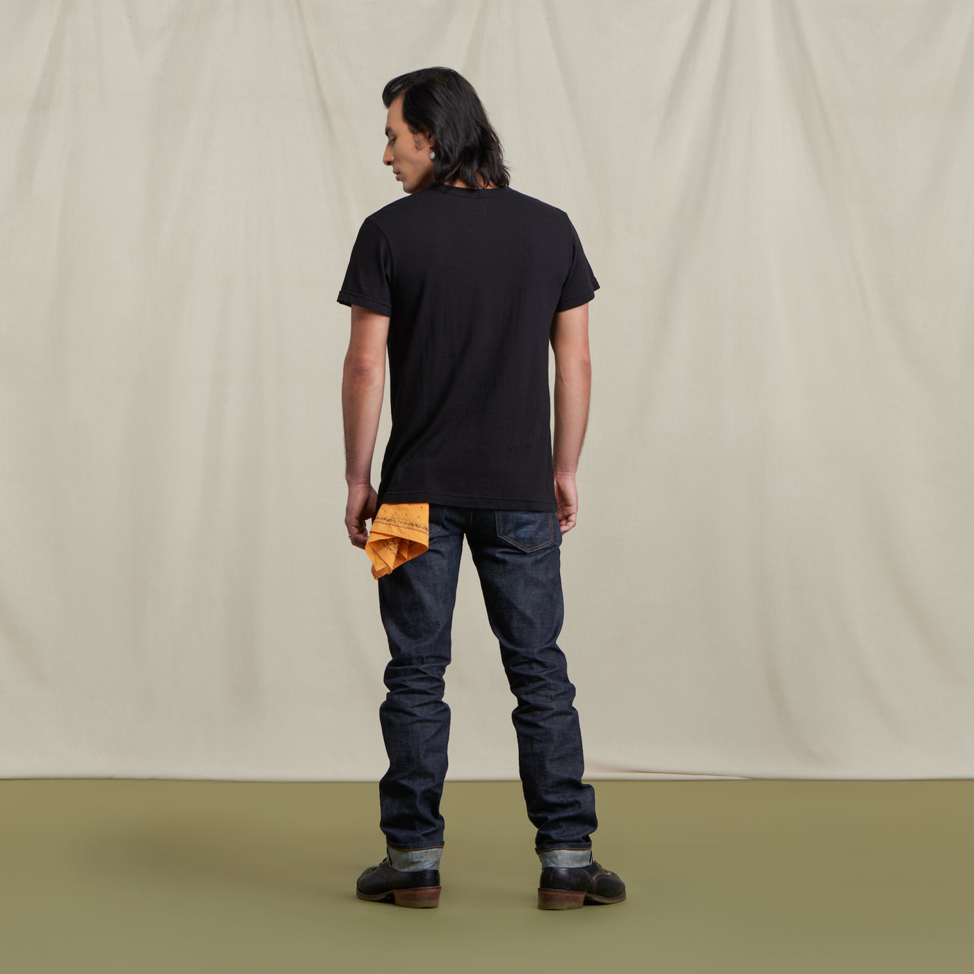The picture is of a male model turned to the back wearing the Crew Tee Jet Black with jeans and brown boots. He is posed in front of a neutral background. He has a yellow bandana in his back pocket.