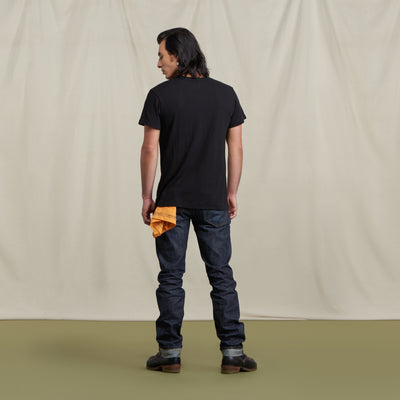 The picture is of a male model turned to the back wearing the Crew Tee Jet Black with jeans and brown boots. He is posed in front of a neutral background. He has a yellow bandana in his back pocket.
