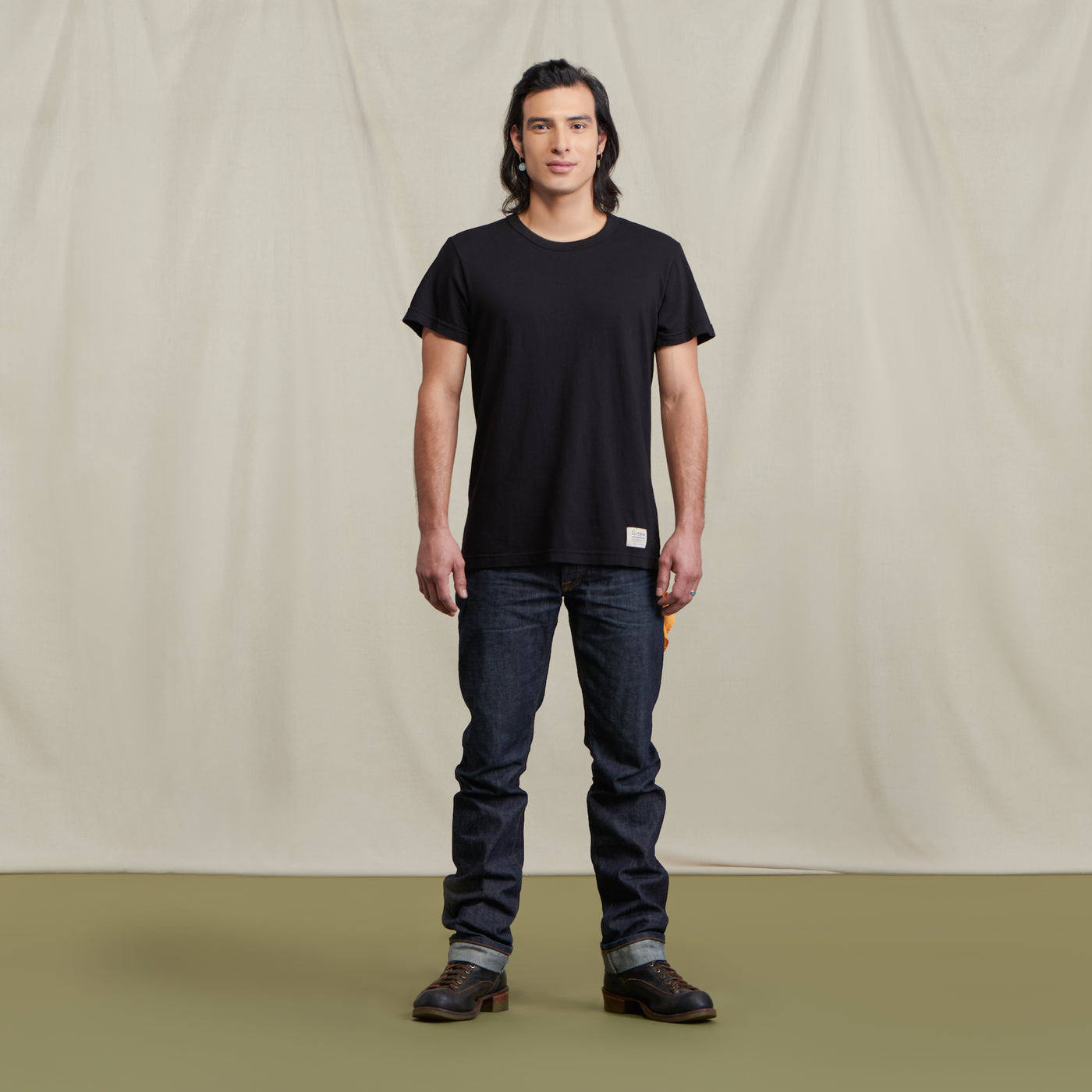 The picture is of a male model wearing the Crew Tee Jet Black with jeans and brown boots. He is posed in front of a neutral background.