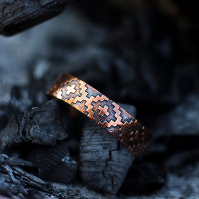 Solid copper Skinny Basket bracelet with a cross design shown on a charcoal log.