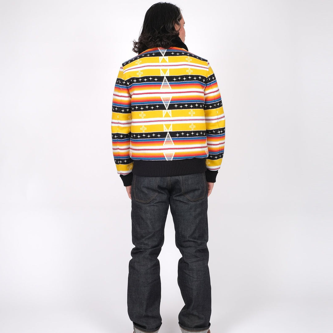 A male model turned to the back wears the yellow and black printed Ginew + Dehen Facing East Flyer's Jacket with jeans and brown shoes against a white background.