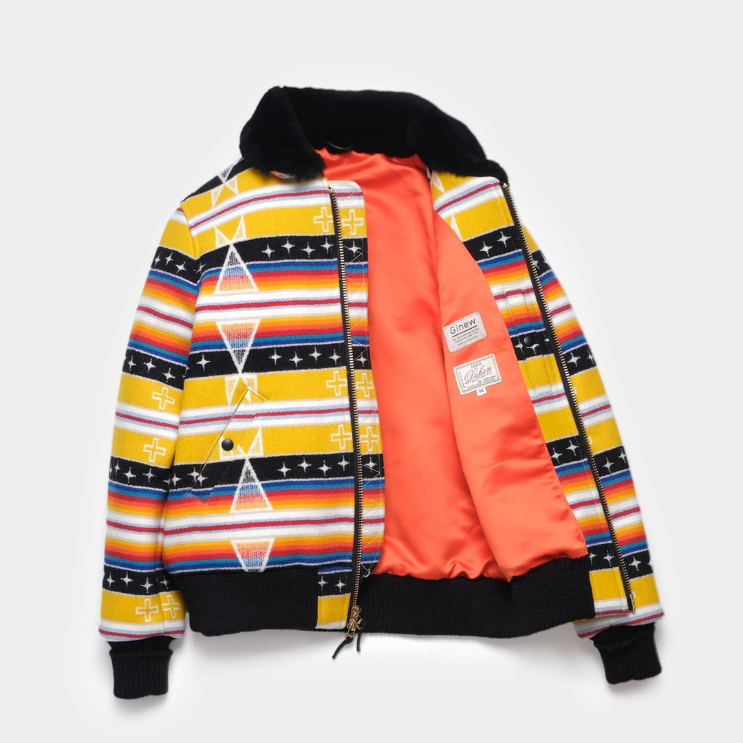 The yellow and black printed Ginew + Dehan Facing East Flyer's Jacket is laid flat on a white background. The jacket is unzipped to show the inner orange lining and white labels from Ginew and Dehen.