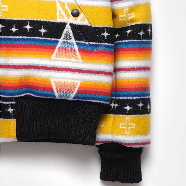 A close-up of the black, ribbed hem and cuffs of the Ginew + Dehen Facing East Flyer's Jacket. The jacket is made from a yellow, black, red, blue, orange, and white patterned wool fabric.