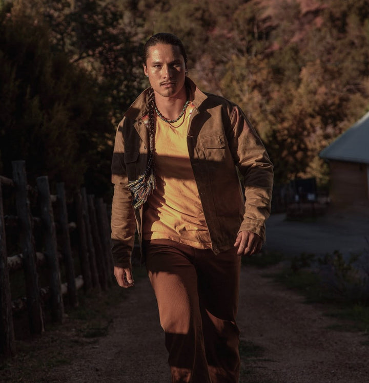 Model wearing Wax Rider Coat in Brown paired with a yellow crew tee and brown pant. Model walking toward camera on a trail.