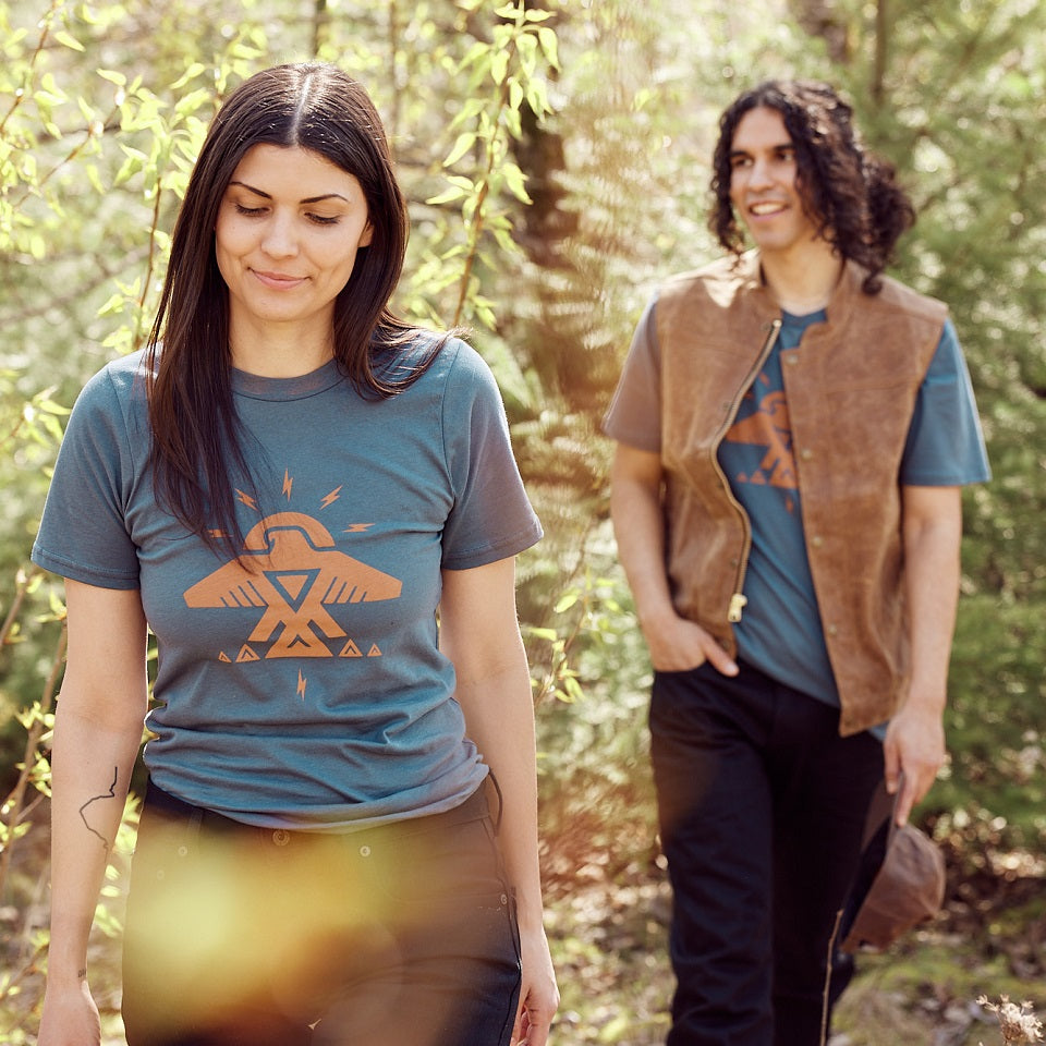 Female and male model wearing tee which is blue with adobe ink graphic of the Ginew Thunderbird, walking toward the camera surrounded by trees.