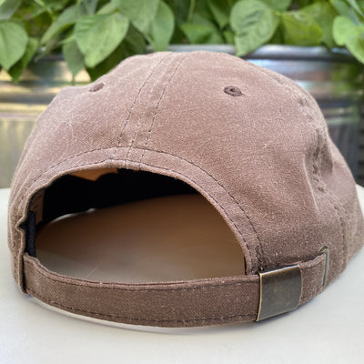 A close-up of the adjustable cloth clamp back of the Thunderbird Patch Ball Cap in a light brown waxed canvas. Shown on a white surface in front of a metal planter with green plants.