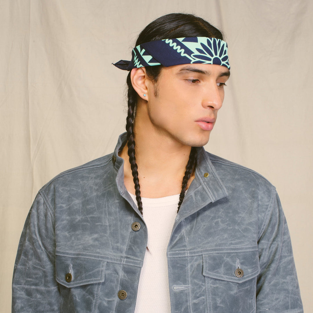 The picture is of a male model wearing the blue and turquoise Naasgo/Forward Movement bandana tied around his head. He is wearing a grey jacket and is posed in front of a neutral background.