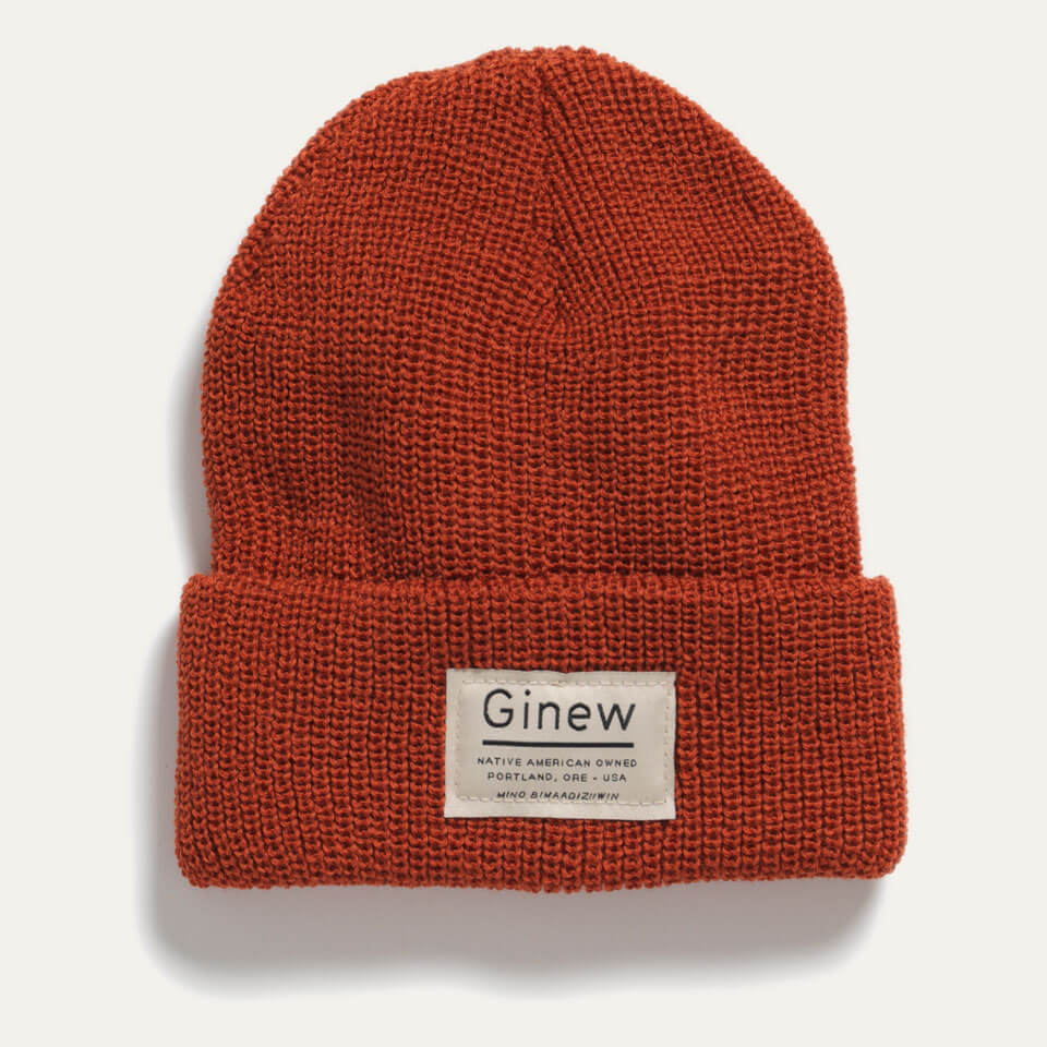 Rust merino wool watch cap beanie shown on a grey background. A white Ginew label that says "Native American Owned Portland, ORE - USA Mino Bimaadiziiwin" is sewed onto the center front.