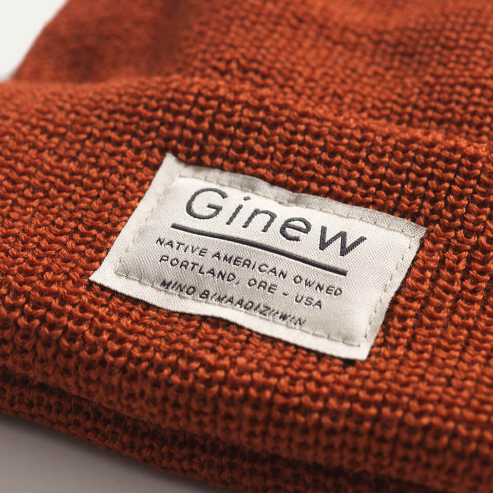 Close-up of a Rust merino wool watch cap beanie shown on a white background. A white Ginew label that says "Native American Owned Portland, ORE - USA Mino Bimaadiziiwin" is sewed onto the center front.