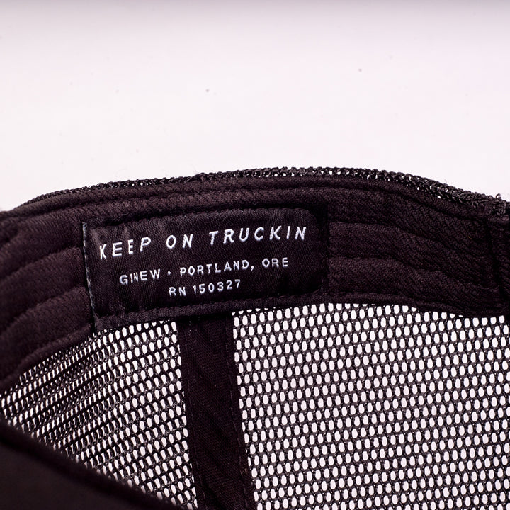A close-up of the inner band of the black Trucker Hat. The photo shows the Ginew tag reading "Keep on Truckin". Shown on a white background.