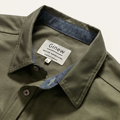 A close-up of an Army green button-up Utility Shirt collar. The shirt has a chambray contrast collar reinforcement with an interior white tag that reads “Ginew, Native America, Mino Bimaadiziiwin”. 