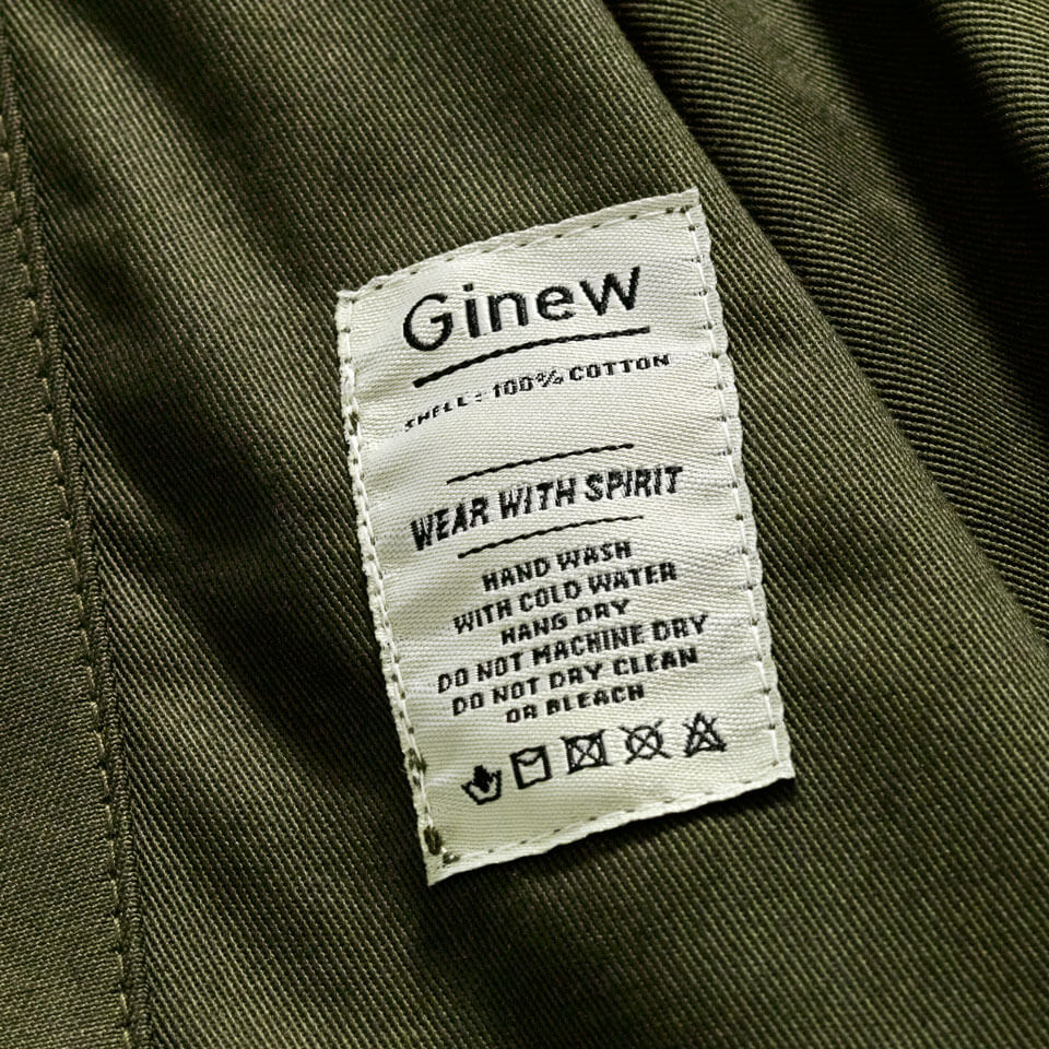 A close-up of an Army green button up Utility Shirt interior white care tag that reads, “Ginew, shell: 100% cotton, Wear with Spirit, hand wash with cold water, hang dry, do not machine dry, do not dry clean or bleach.”