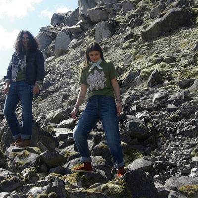 Two models wear water remembers tee. Both models wear jeans and are walking over rocks on a hillside. Male model wearing Thunderbird Jacket over the top. Women wears Water Remembers bandana around her neck.