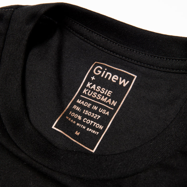 Close-up of the printed neck label of the black Diamond Basket tee. The label is printed in light brown ink and reads "Ginew + Kassie Kussman. Made in USA. RN: 150327. 100% cotton. Wear with spirit. Size M". The shirt is laid on a white background.