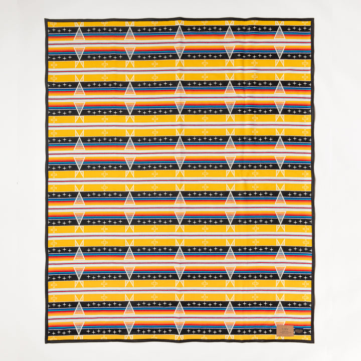 The Facing East wool blanket is yellow, black, white, red, and blue and features a diamond pattern. Shown laid flat against a white background. Native American designed. Ginew: Native American-owned.