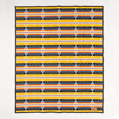 The Facing East wool blanket is yellow, black, white, red, and blue and features a diamond pattern. Shown laid flat against a white background. Native American designed. Ginew: Native American-owned.