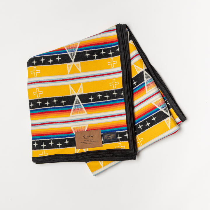 A folded Facing East wool blanket is set against a white background. The wool blanket has a diamond pattern and is yellow, black, white, red, orange, and blue. Native artist designed. Ginew: Native American Owned. 