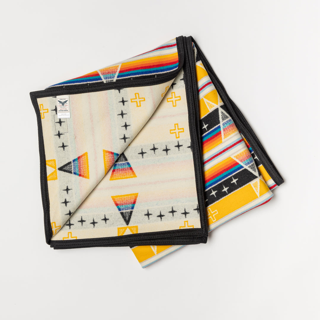 A folded Facing East wool blanket is set against a white background. The wool blanket has a diamond pattern and is yellow, black, white, red, orange, and blue. The blanket is showing the contrasting interior pattern.