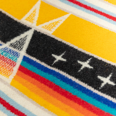 A close-up of the Facing East wool blanket. The blanket is yellow, black, white, red, and blue and features a diamond pattern.