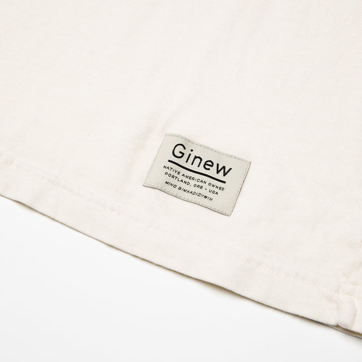 Close-up of a white tag on the bottom left hem on the Crew Tee in Arctic Wolf. The picture shows the white Ginew tag on the bottom left hem which reads "Native American Owned Portland, ORE - USA Mino Bimaadiziiwin". The shirt is laid flat on a white background.