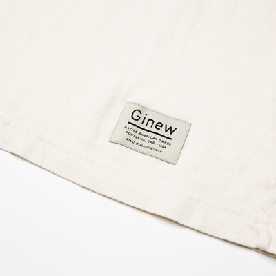 Close-up of a white tag on the bottom left hem on the Crew Tee in Arctic Wolf. The picture shows the white Ginew tag on the bottom left hem which reads "Native American Owned Portland, ORE - USA Mino Bimaadiziiwin". The shirt is laid flat on a white background.