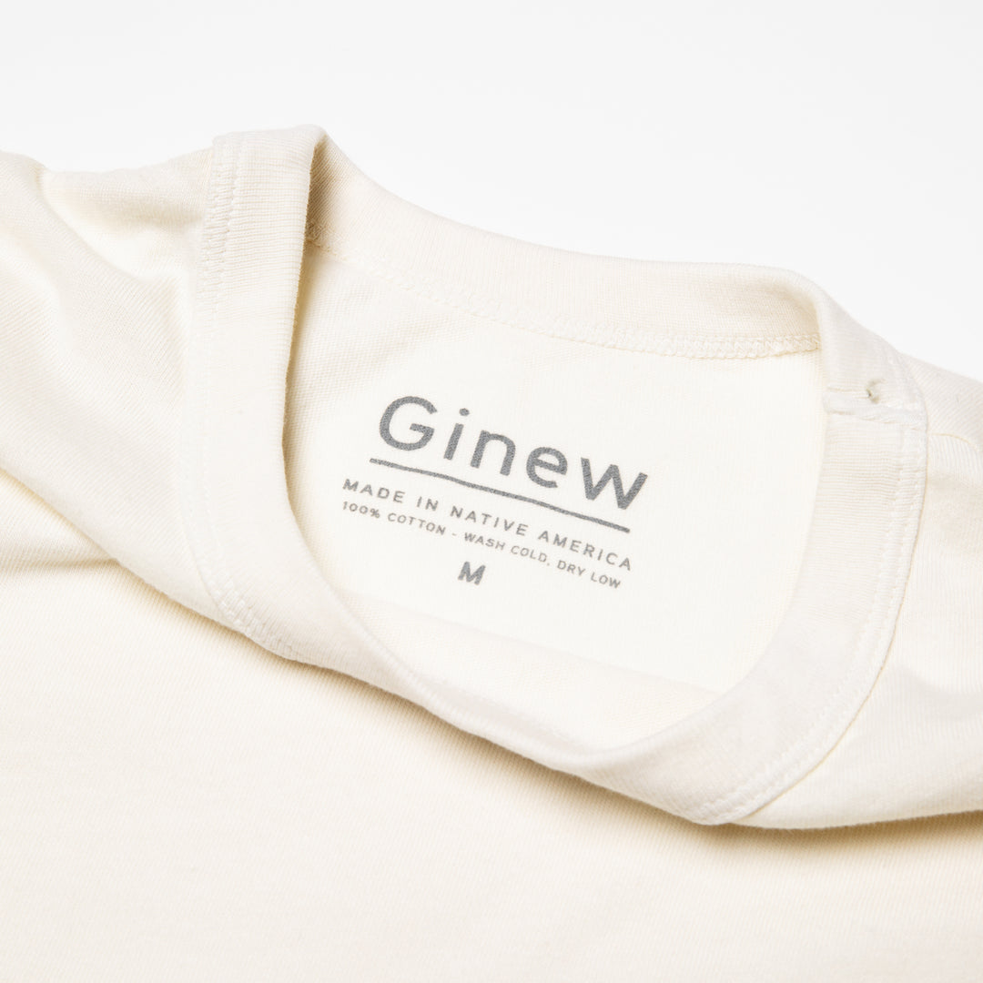 Close-up of the back neck label on white tshirt. The label is printed on the shirt in grey ink. The label says "Ginew. Made in Native America. 100% cotton. 