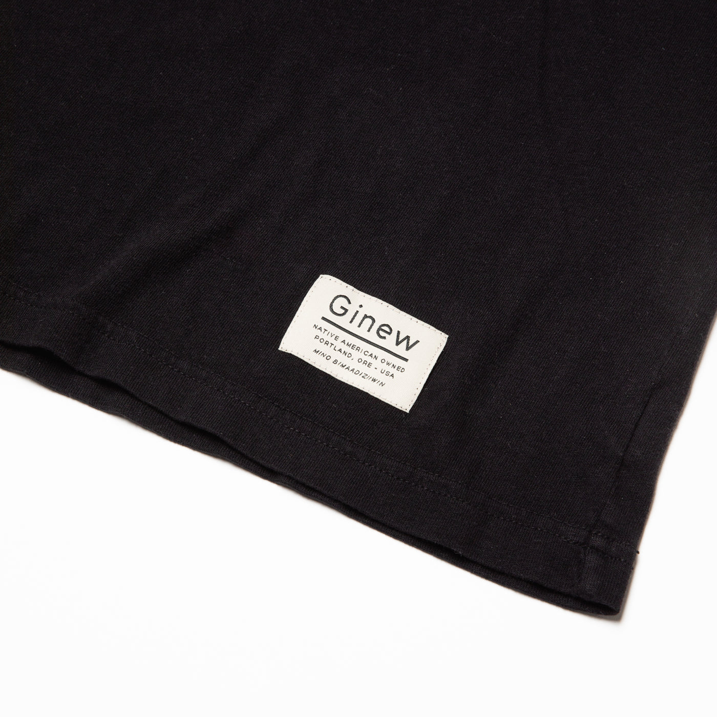The Crew Tee in Jet Black. The picture shows the white Ginew tag on the bottom left hem which reads "Native American Owned Portland, ORE - USA Mino Bimaadiziiwin". The shirt is laid flat on a white background.