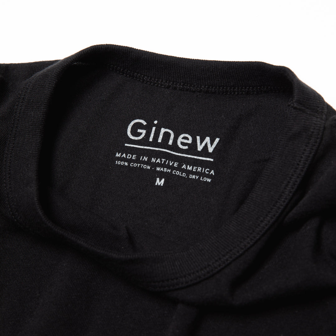 Close-up of the back neck label of the Crew Tee in Jet Black. The label is printed on the shirt in grey ink. The label says "Ginew. Made in Native America. 100% cotton. Wash cold, dry low. Size M". The shirt is laid on a white backgound.