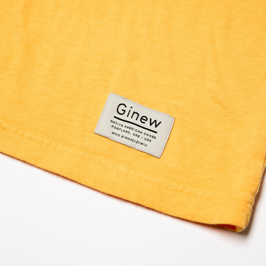 Close-up of a white tag on the bottom left hem on the Crew Tee in Marigold yellow. The picture shows the white Ginew tag on the bottom left hem which reads "Native American Owned Portland, ORE - USA Mino Bimaadiziiwin". The shirt is laid flat on a white background.