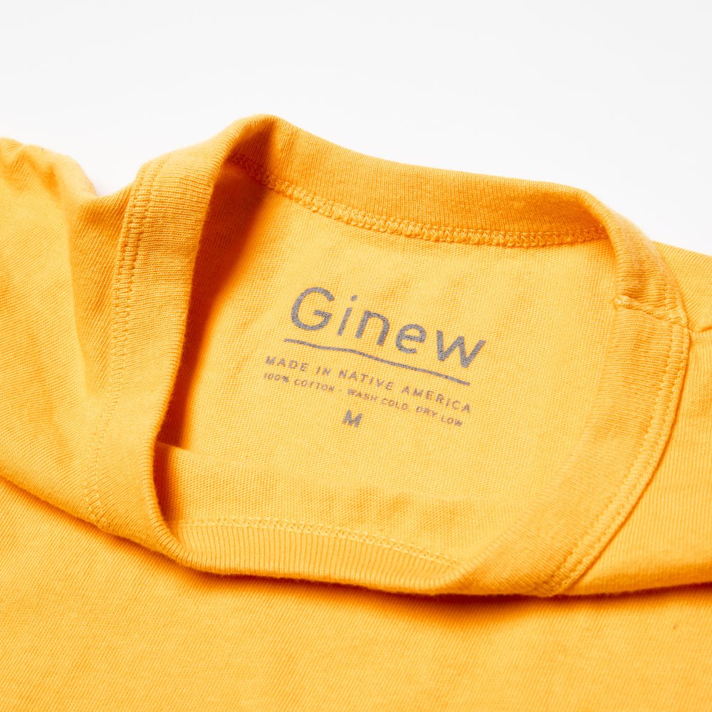 Close-up of the back neck label of the Crew Tee in Marigold. The label is printed on the shirt in grey ink. The label says "Ginew. Made in Native America. 100% cotton. Wash cold, dry low. Size M". The shirt is laid on a white backgound.