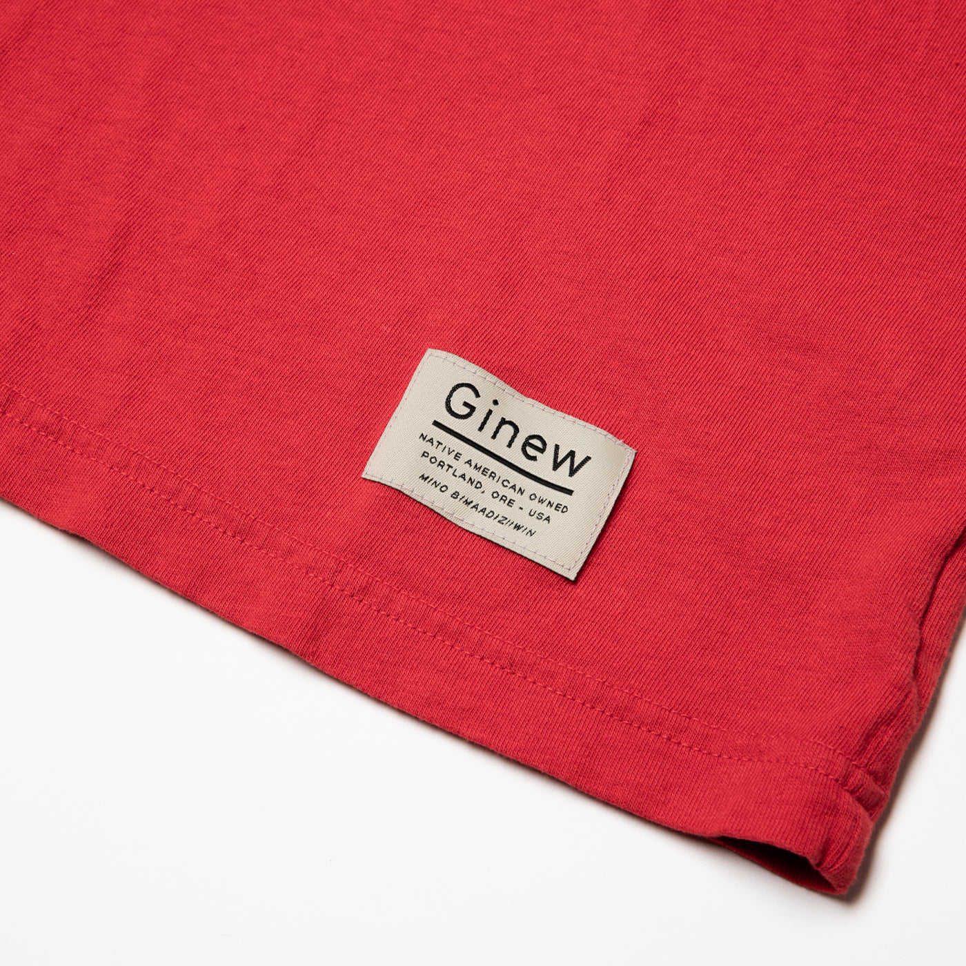 Close-up of a white tag on the bottom left hem on the Crew Tee in Urban Red. The picture shows the white Ginew tag on the bottom left hem which reads "Native American Owned Portland, ORE - USA Mino Bimaadiziiwin". The shirt is laid flat on a white background.