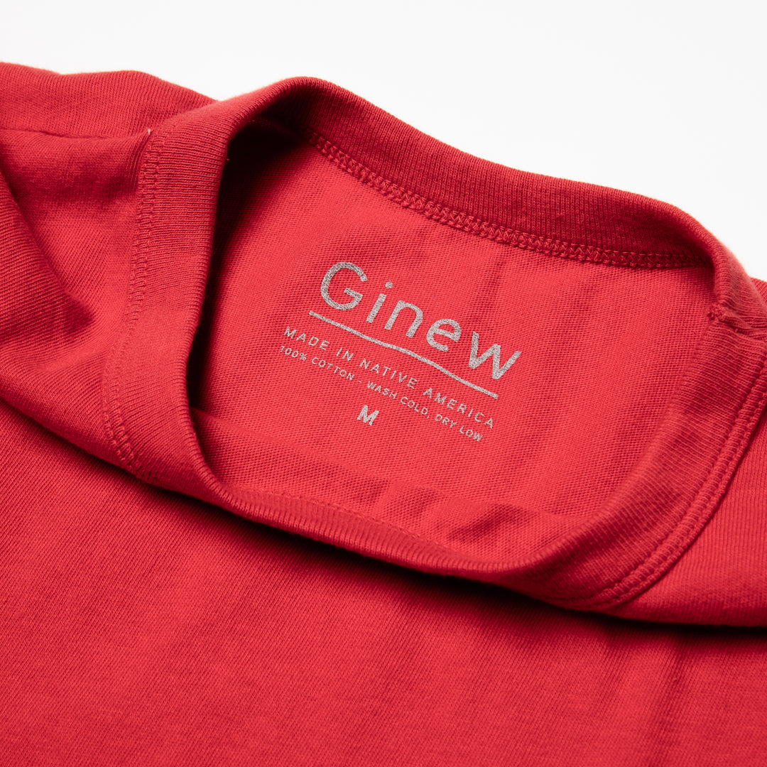 Close-up of the back neck label of the Crew Tee in Urban Red. The label is printed on the shirt in grey ink. The label says "Ginew. Made in Native America. 100% cotton. Wash cold, dry low. Size M". The shirt is laid on a white backgound.