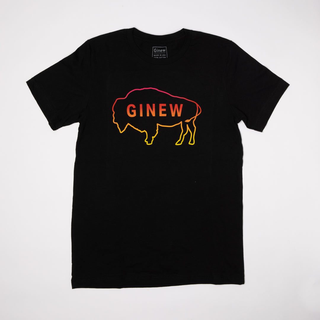 A black t-shirt lays on a wood floor. The t-shirt has an outline of a buffalo with the word Ginew in the center. The colors of the buffalo and word start with a neon pink at the top and fade through red and orange to yellow.