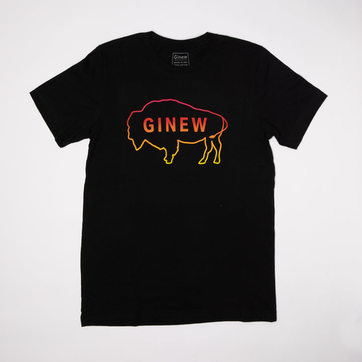 A black t-shirt lays on a wood floor. The t-shirt has an outline of a buffalo with the word Ginew in the center. The colors of the buffalo and word start with a neon pink at the top and fade through red and orange to yellow.