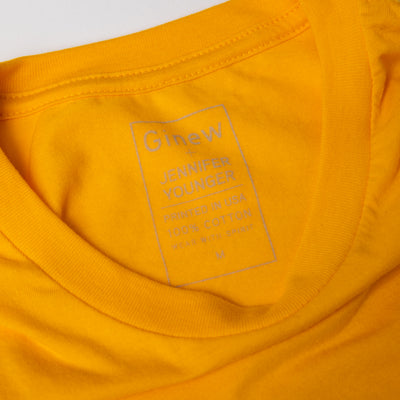 Close up of inside of back of tee with printed tag that reads: "GINEW - JENNIFER YOUNGER - PRINTED IN USA - 100% COTTON - WEAR WITH SPIRIT"