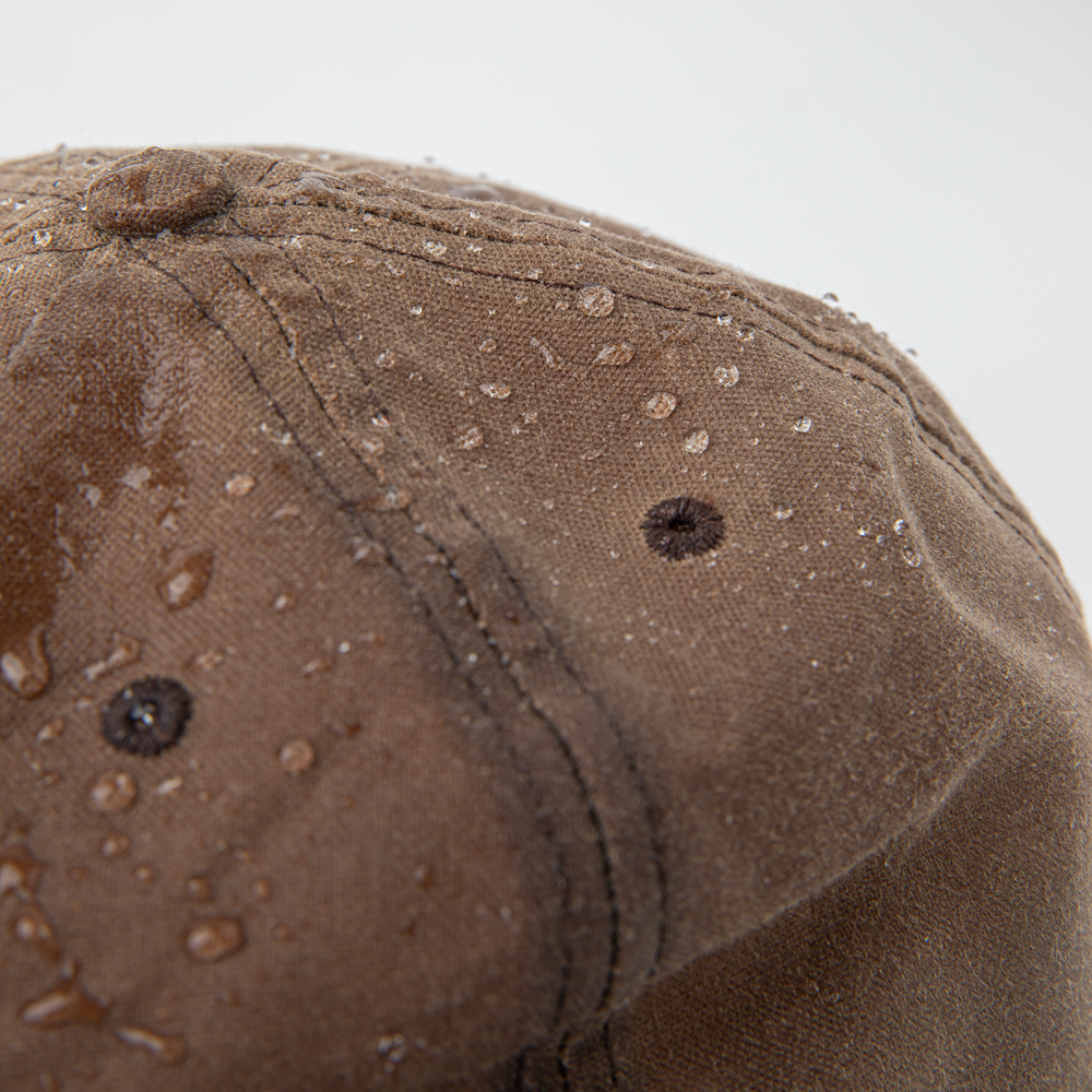 A close up of the top of the light brown waxed canvas Crow Wing Ball Cap showing the water-repellant quality with beads of water on the outside of the cap. Shown on a white background.