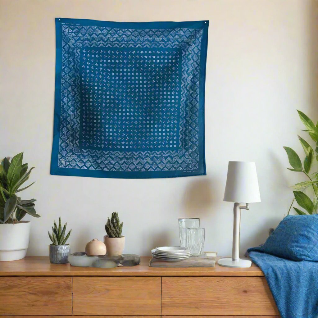 The Indigo Medel Bandana hangs on a wall of house next to a framed picture and above a chair and a table with books and a water bottle.