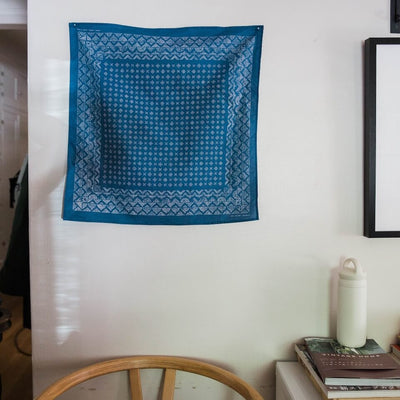 The Indigo Medel Bandana hangs on a wall of house next to a framed picture and above a chair and a table with books and a water bottle.